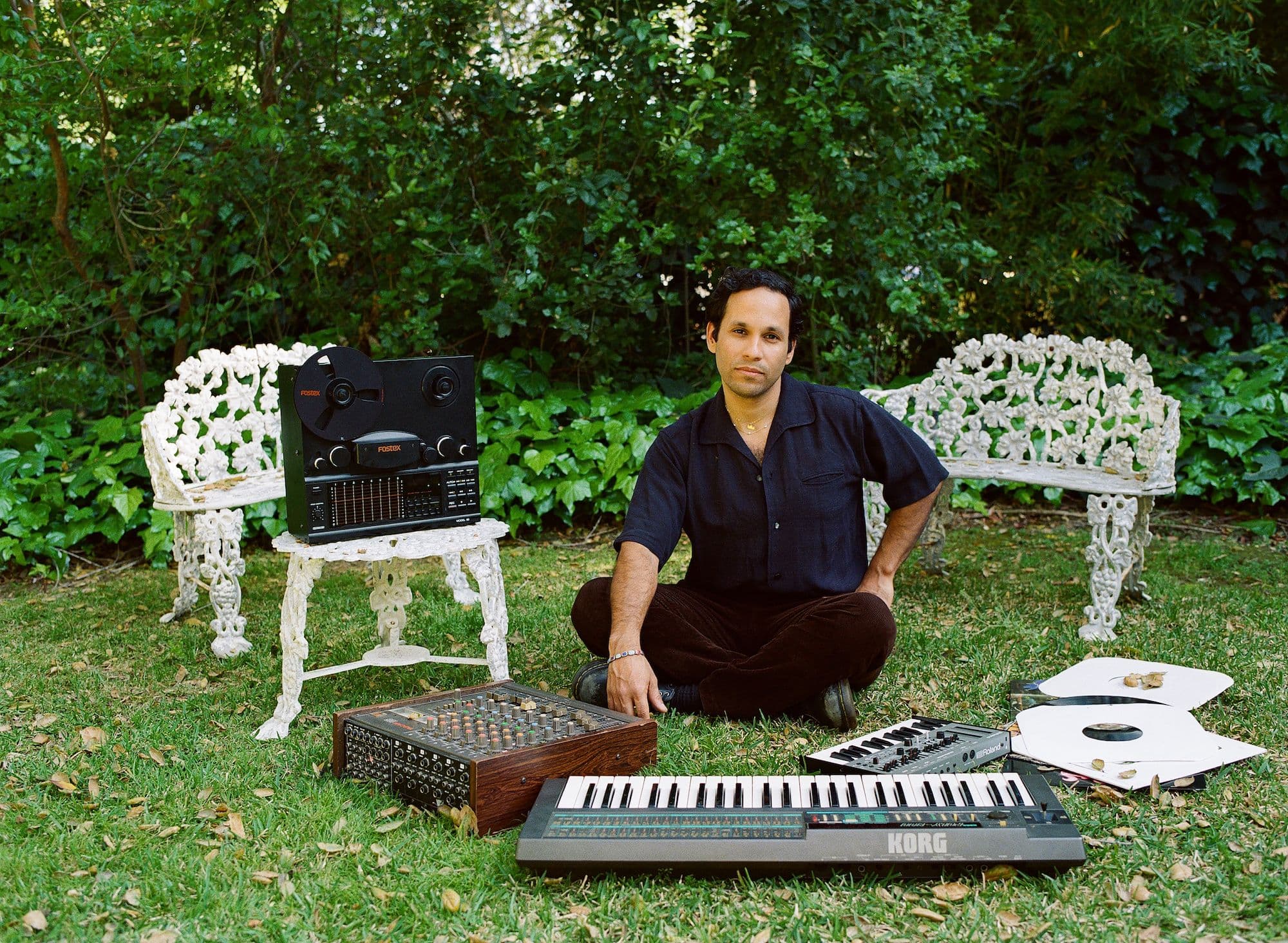A man is seated on the grass, using a keyboard and electronic gear, immersed in his creative process.