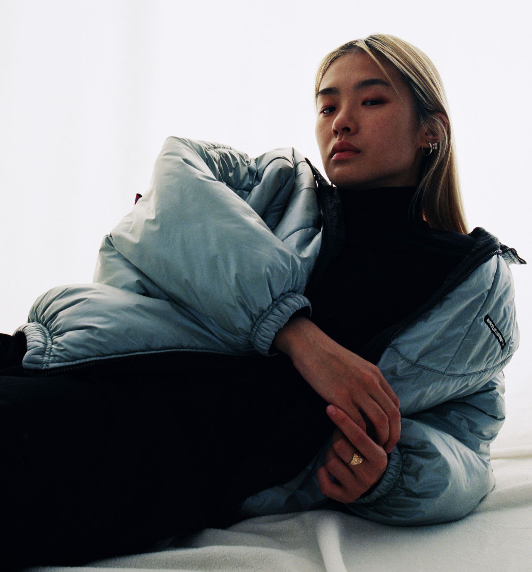 A woman in a blue puffy jacket is relaxing on a bed, looking comfortable and cozy.